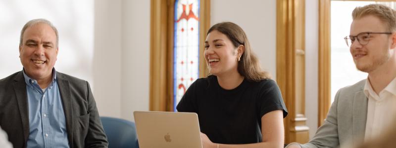 Three people smiling around a table, one with a laptop, in a well-lit room.