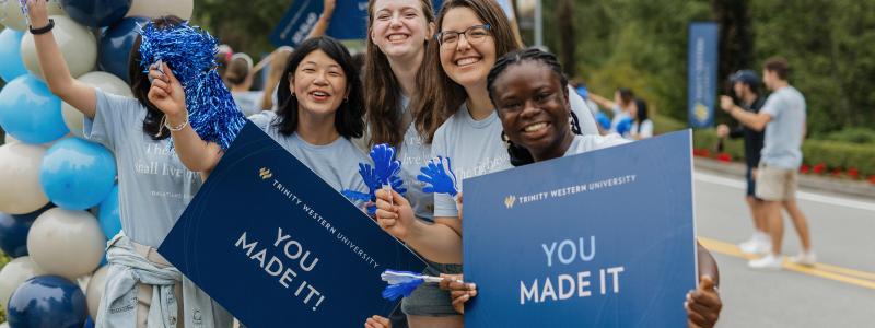 Four people smiling, holding blue signs saying "YOU MADE IT" with balloons nearby.