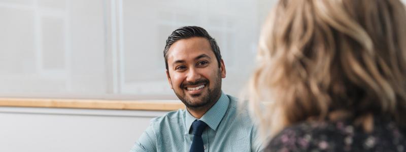 Man in a tie smiling, talking to a woman in an office setting.