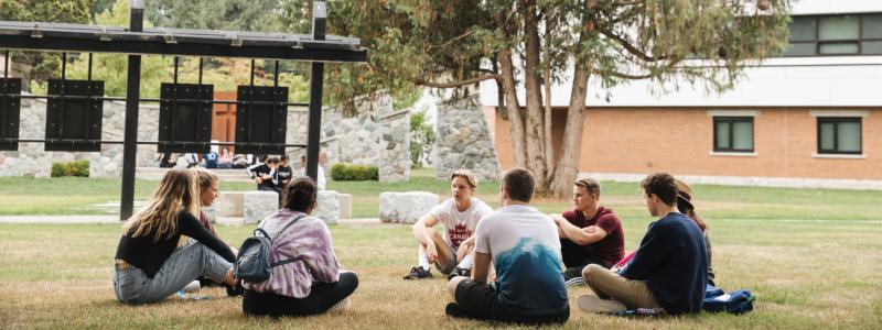 Students sitting in a circle on grass, school building in background.