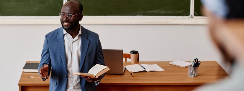 Man in blue blazer teaching in a classroom with a chalkboard.