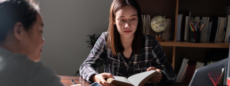 Two people study together at a table with a book and laptop.