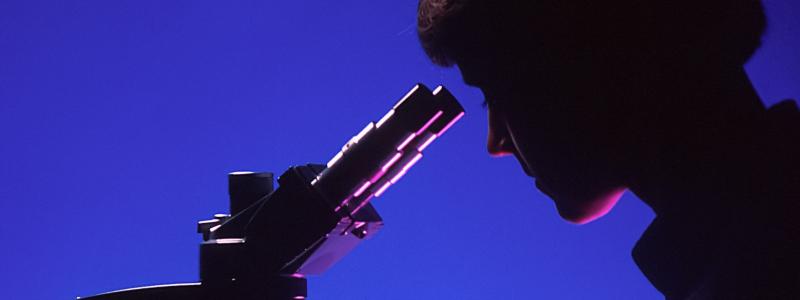 Silhouette of person using a microscope against a blue background.