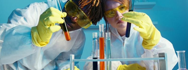 Two scientists in lab coats examine test tubes with serious expressions.
