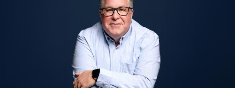 Smiling man with glasses in a blue shirt against a dark blue background.