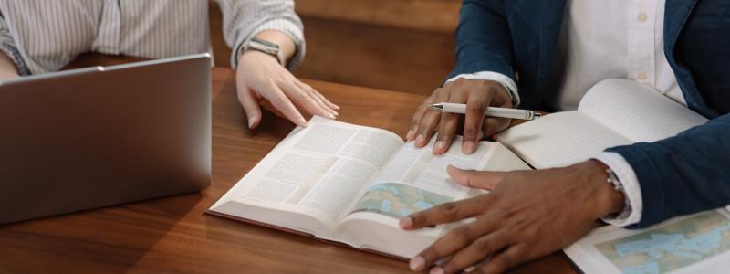Two people studying a Bible and using a laptop at a wooden table.