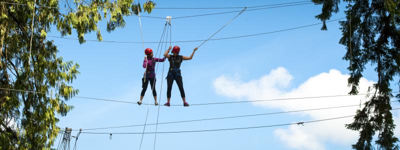 Two people on a rope course in a forest, wearing helmets, under a blue sky.