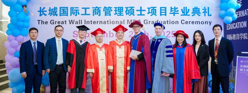 Graduates and officials in robes pose at a ceremony, with blue balloons and a banner backdrop.
