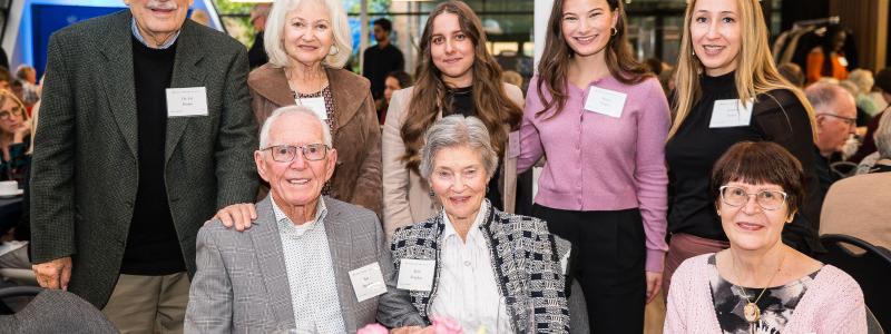 Group of people of various ages smiling at an event, gathered around a table with flowers.