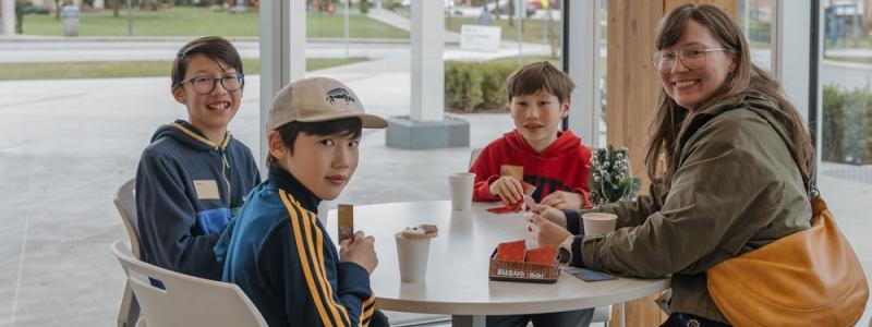 Woman and three young boys sitting at a table with paper cup drinks, playing cards, facing the camera with a smile.