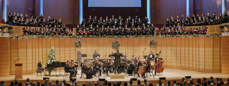 A full orchestra performing on stage and a conductor leading them during the Christmas concert. Behind the orchestra, a large choir stands on an elevated platform, and the stage is decorated with wreaths, garlands, and a Christmas tree, creating a festive atmosphere.