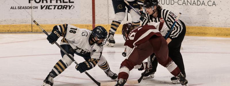 Two hockey players face off on the ice, one in a white TWU jersey and the other in a maroon uniform, with a referee dropping the puck and another player in white positioned behind them. Advertising boards are visible in the background.