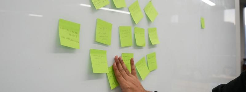 A person places or adjusts bright green sticky notes on a whiteboard, which is covered with several handwritten notes arranged in a cluster.