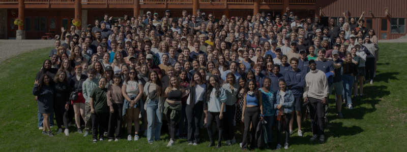 A large group of students stand outside with a wooden building in the background.