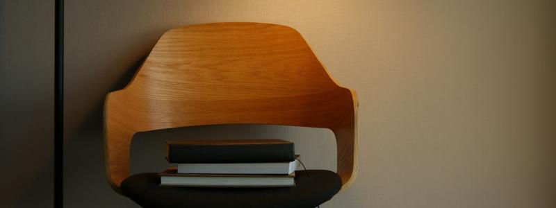 A wooden chair with a black cushioned seat sits against a neutral wall, illuminated by soft lighting. A small stack of books rests neatly on the seat.