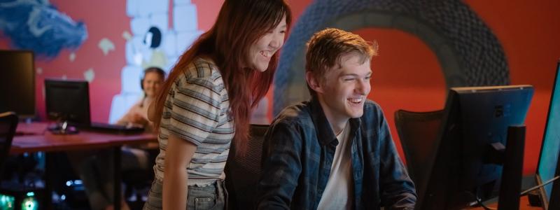 Two people work together at a computer station in a dimly lit room with a colorful mural on the wall. 