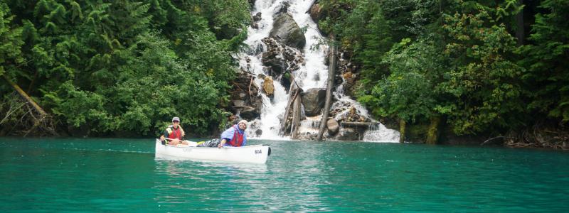 Two people canoe on turquoise water in front of a cascading waterfall surrounded by dense green forest.