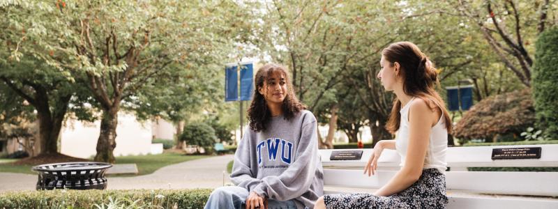 Two student sitting outdoors on a bench, surrounded by green trees.