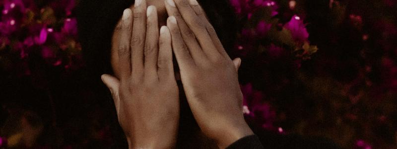 Man covering his face with his hands with a bright pink bush of flowers in the background