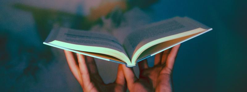 Two hands hold an open book in midair against a softly lit, colorful background, with the pages glowing slightly from the light.