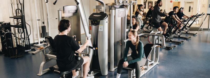 People exercise in a bright fitness center, using strength machines and stationary bikes, with others resting or talking between workouts.