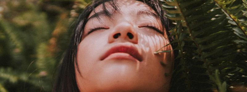 A woman with long dark hair is positioned outdoors among dense, green fern leaves. 