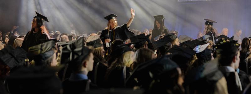 graduate walking in a cap and gown in a room with spotlights and the audience around her
