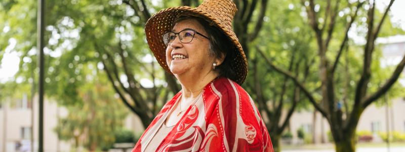 Patti Victor Switametelót in her cultural regalia smiling and looking past the camera in front of greenery