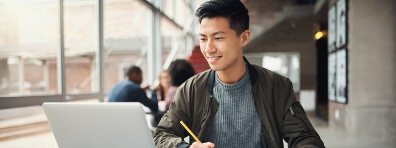 image of a person looking at the laptop, holding a pen and has a book infront of him 