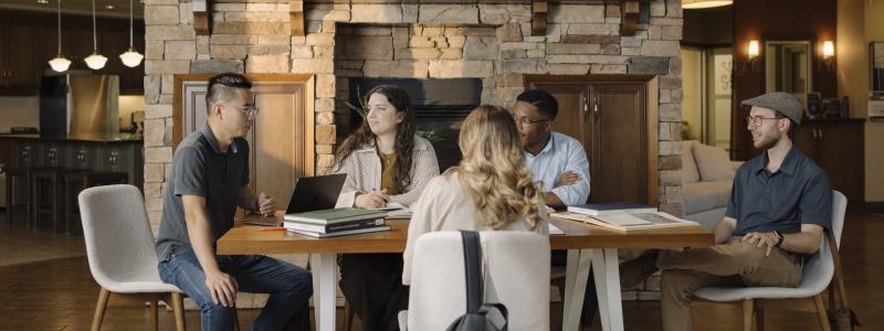 TEDS students gather around a table in the Graduate Collegium, participating in a discussion with laptops, books, and notebooks in a lounge-style learning space featuring a large stone fireplace.