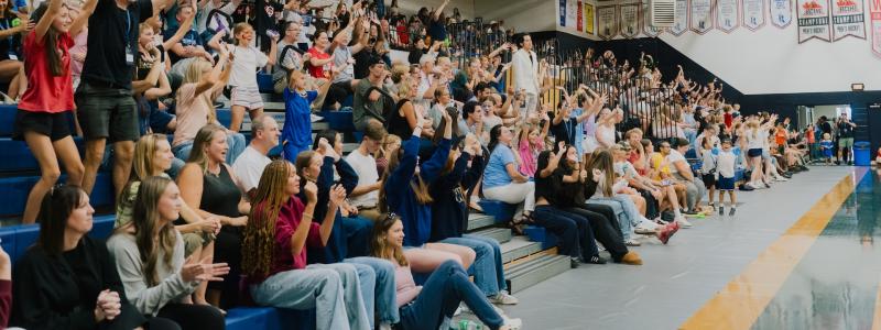 A large crowd of spectators fills the bleachers inside a university gymnasium, with many people raising their hands and cheering during a campus event. Championship banners hang on the wall above the court, and bright overhead lights illuminate the packed stands.