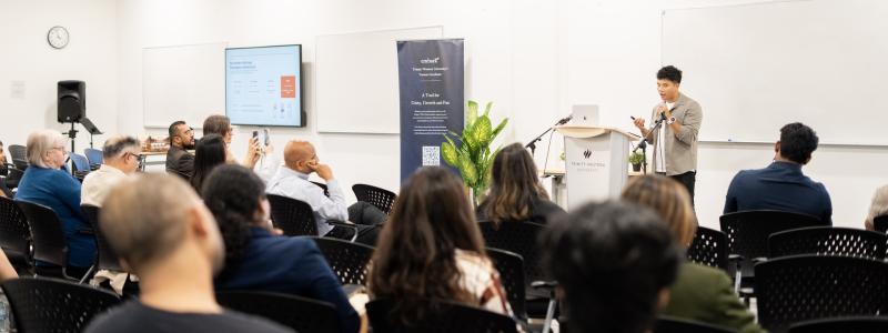 Audience members seated in a classroom-style event space at TWU Richmond campus listen as a speaker presents from a podium. A presentation screen, TWU-branded banner, and whiteboards are visible at the front of the room.