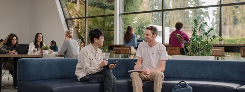 Students connect and study in Trinity Commons on Trinity Western University's Langley campus, using collaborative seating and workspaces beside large windows overlooking the campus landscape.