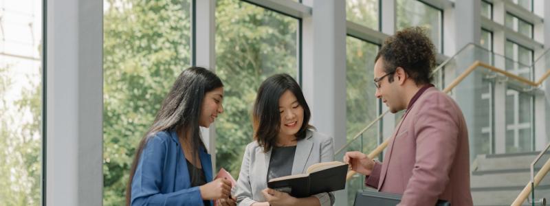 Three people in business attire look at an open notebook together beside large windows and a staircase at TWU.