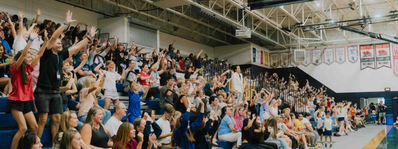 A crowd of adults and children cheer with raised hands from gymnasium bleachers during TWU Homecoming.