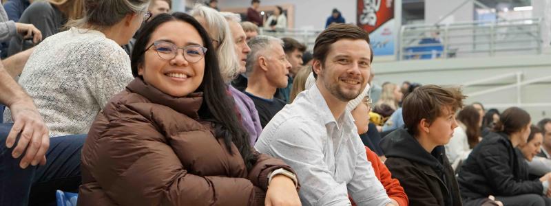 Two attendees smiling at the camera while seated among spectators in gymnasium.