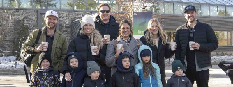 A group of adults and children dressed for cool weather gather outdoors in front of a campus building while holding warm drinks.