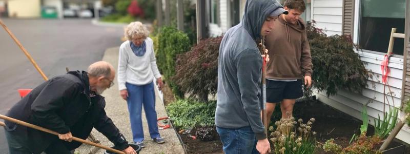 group of people working together outdoors on a community service project, using rakes and shovels around a garden area.