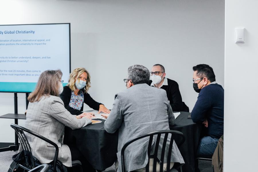 Staff members gathered around a table discussing. There is a PowerPoint in the background.