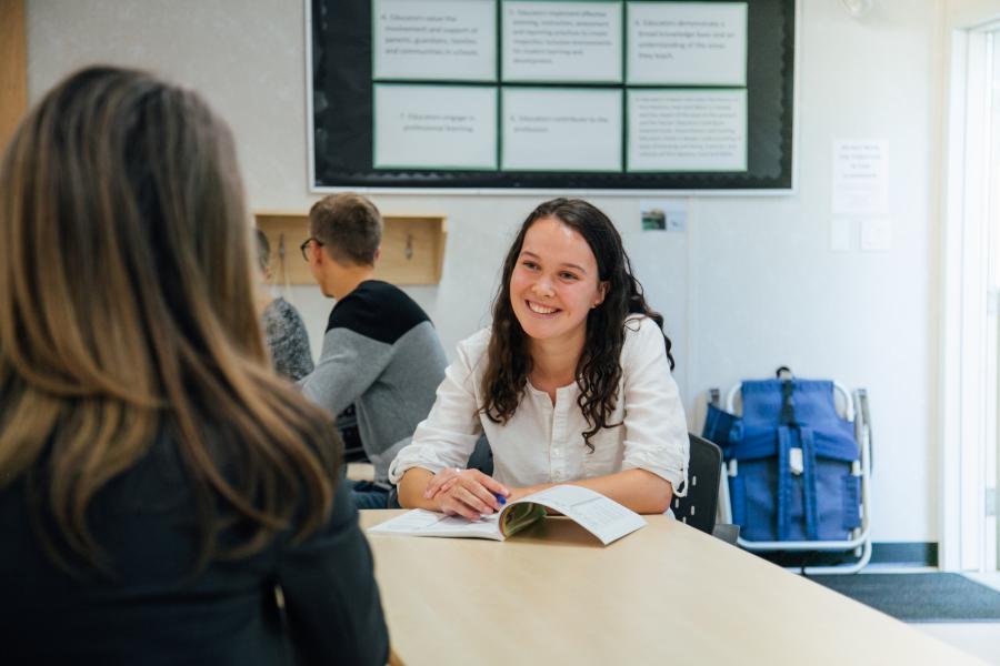 Female student at a desk, smiling at her classmate, with a booklet open.