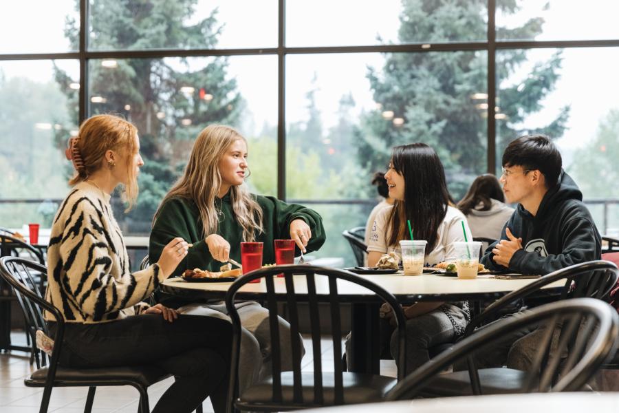 Four students sitting in cafeteria eating, smiling, talking.