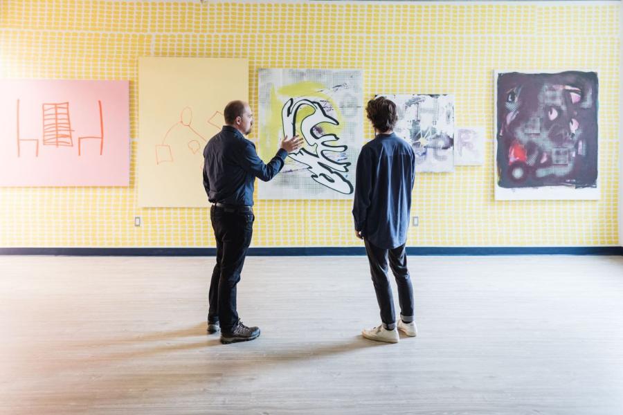 A professor and a student talking while looking at a wall displaying five art pieces.