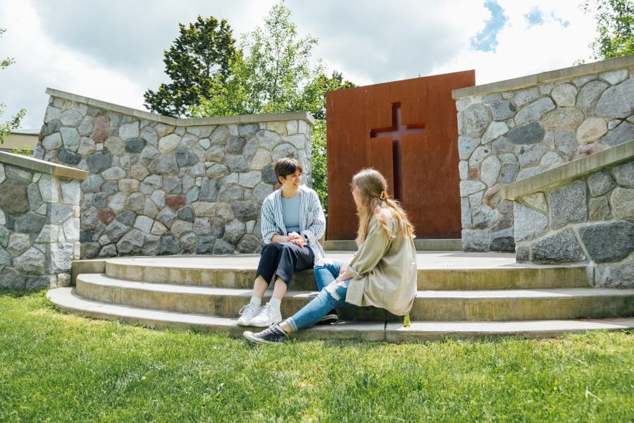Two female students sitting on the steps of the Langley campus outdoor chapel talking. There is a cross in the background.