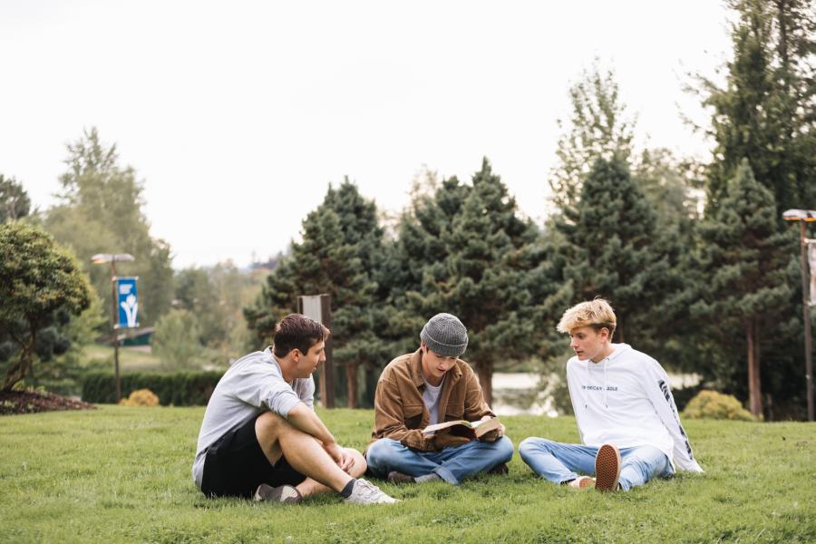 Three male students sitting on grass at the TWU Langley campus reading the bible and talking.