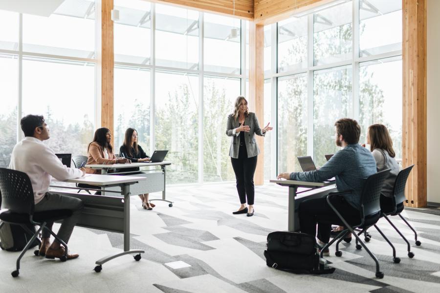 Teacher speaking to students sitting in a classroom with windows