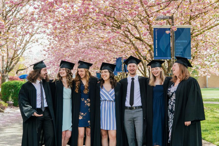 Seven students in graduation cap and gown smiling under the cherry blossoms with their arms around each other.