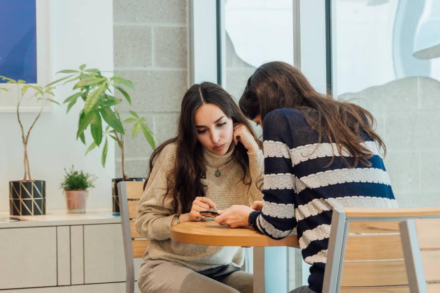 two students sitting at table and looking at phone together