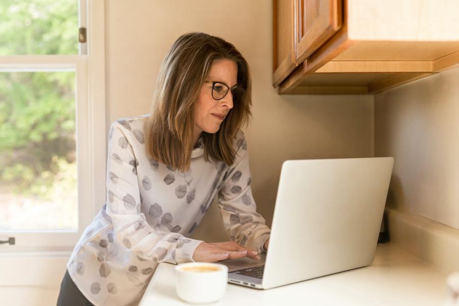 Woman in kitchen working on laptop while standing at a cupboard