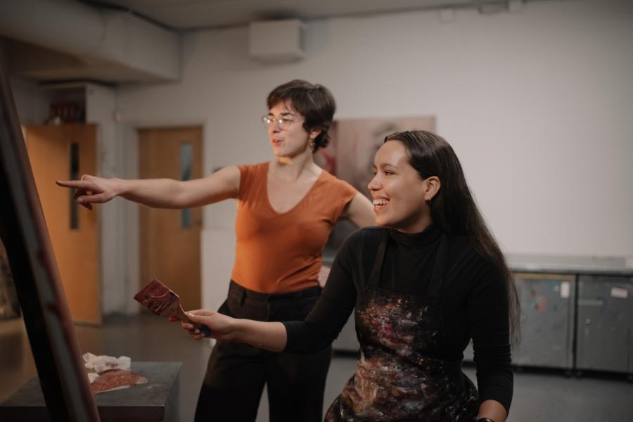 A student holding a paintbrush, with an instructor pointing at her work and smiling