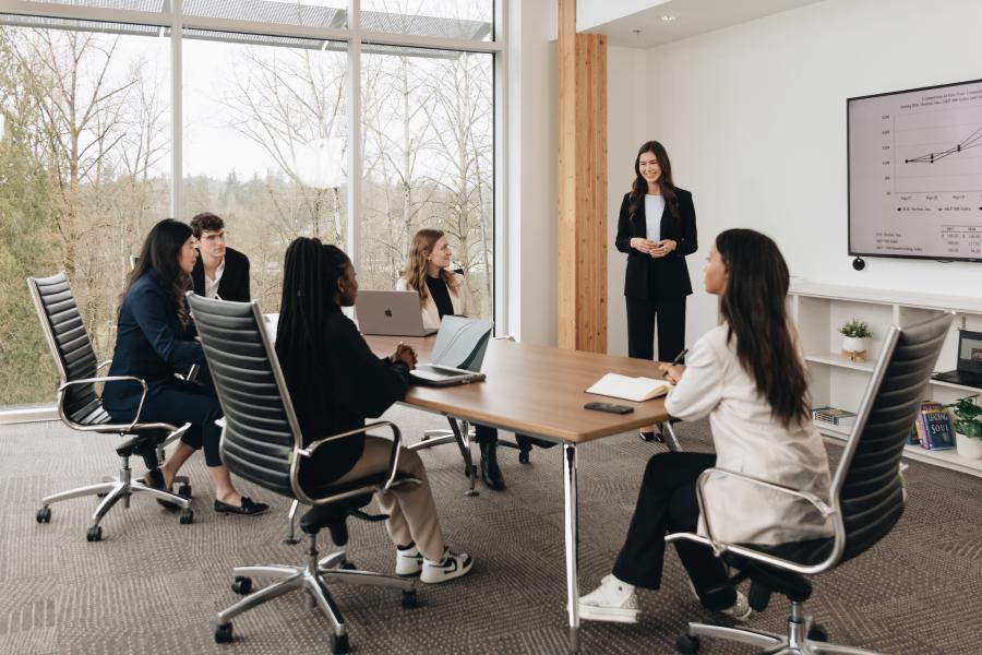 A professional meeting is taking place in a modern conference room with floor-to-ceiling windows overlooking a wooded landscape. A woman in a black suit is standing at the front of the room, presenting a line graph displayed on a large screen. Five colleagues, seated around a wooden table, are attentively listening, taking notes, and using laptops. The room features sleek black office chairs, built-in shelving with books and plants, and natural wood accents, creating a bright and contemporary workspace.
